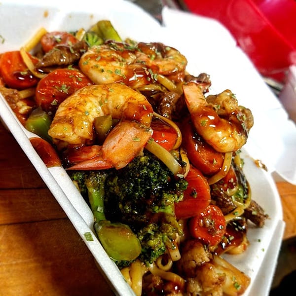 Cajun shrimp and beef stir-fry with broccoli, tomatoes, and noodles in a white takeout container.